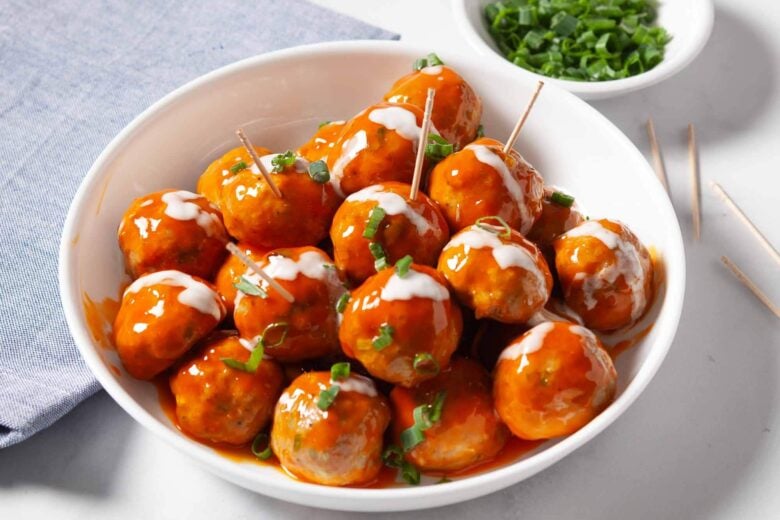 A white bowl filled with buffalo chicken meatballs, glazed and garnished with chopped green onions and a drizzle of ranch dressing, served as spicy appetizers with toothpicks inserted in some meatballs.