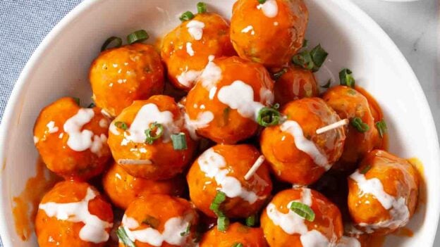 A white plate of buffalo chicken meatballs coated in spicy honey hot sauce, drizzled with white dressing and topped with chopped green onions, sits next to a blue napkin and a small bowl of green onions.