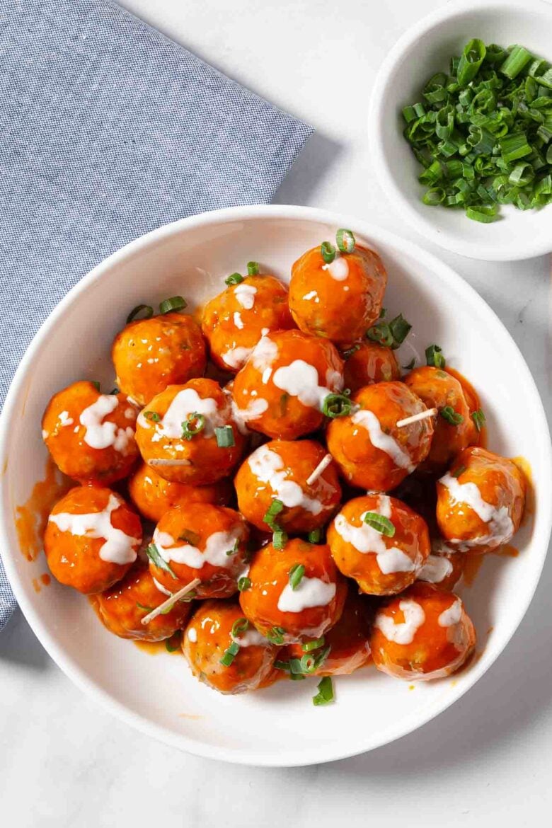 A white plate of buffalo chicken meatballs coated in spicy honey hot sauce, drizzled with white dressing and topped with chopped green onions, sits next to a blue napkin and a small bowl of green onions.