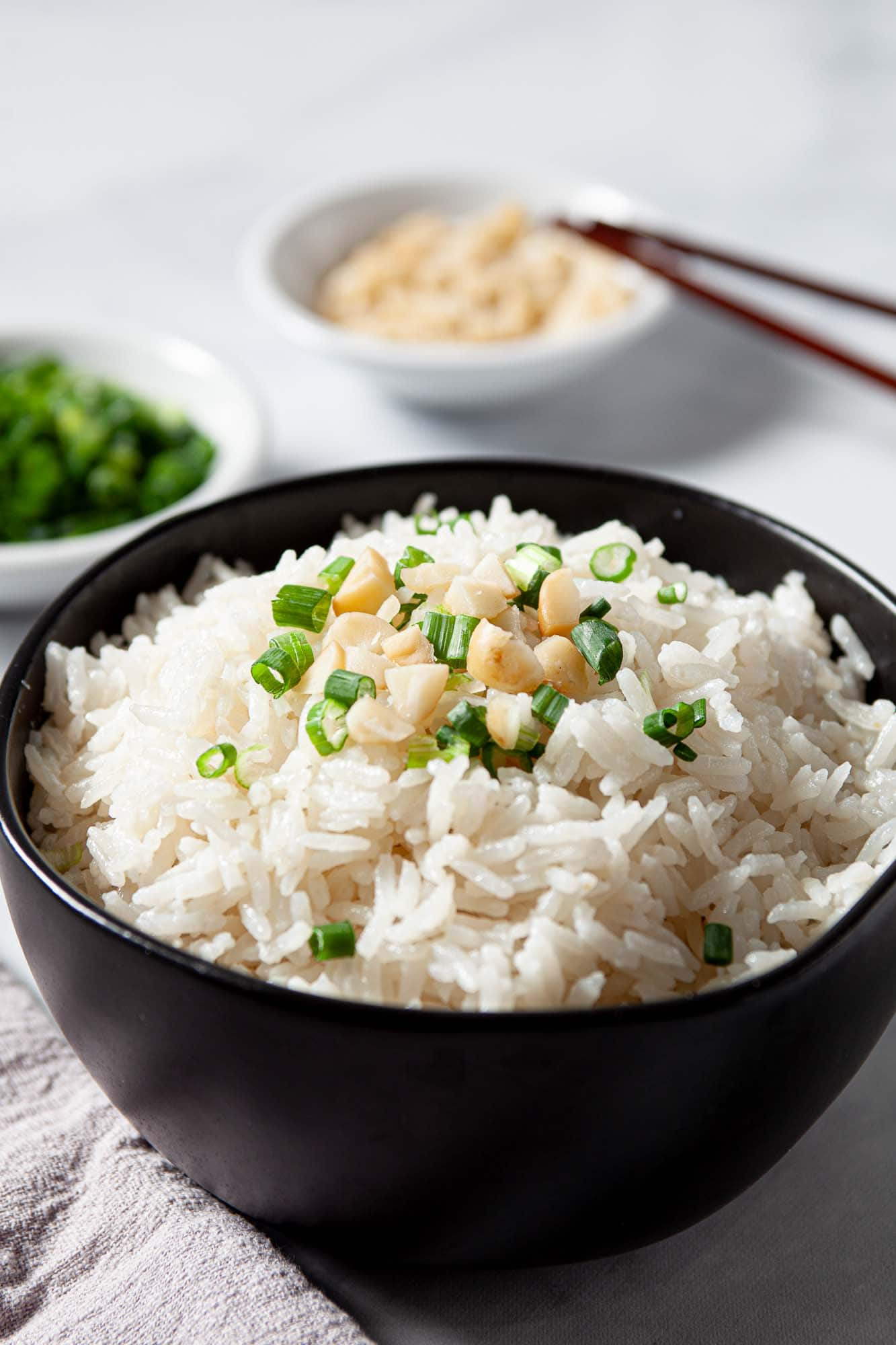 A black bowl filled with tropical coconut rice, topped with chopped green onions and garlic, sits on a table with small dishes of more green onions and garlic in the background.