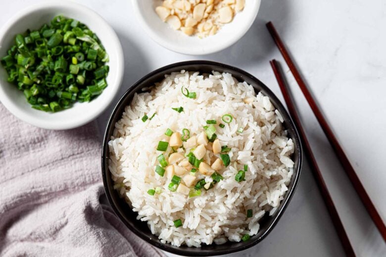 A bowl of Asian style coconut rice topped with chopped green onions and peanuts, next to chopsticks, a bowl of green onions, and a bowl of chopped peanuts on a white surface.