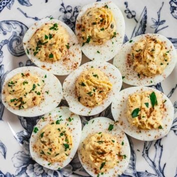 A plate with eight creamy deviled eggs, topped with paprika and chopped herbs, arranged neatly on a decorative blue and white dish&mdash;an ideal make-ahead appetizer for any gathering.