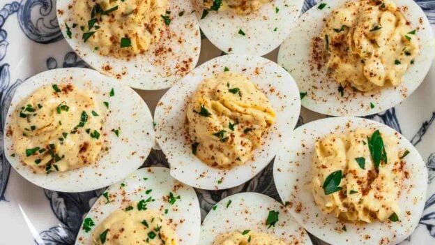 A plate with eight creamy deviled eggs, topped with paprika and chopped herbs, arranged neatly on a decorative blue and white dish&mdash;an ideal make-ahead appetizer for any gathering.