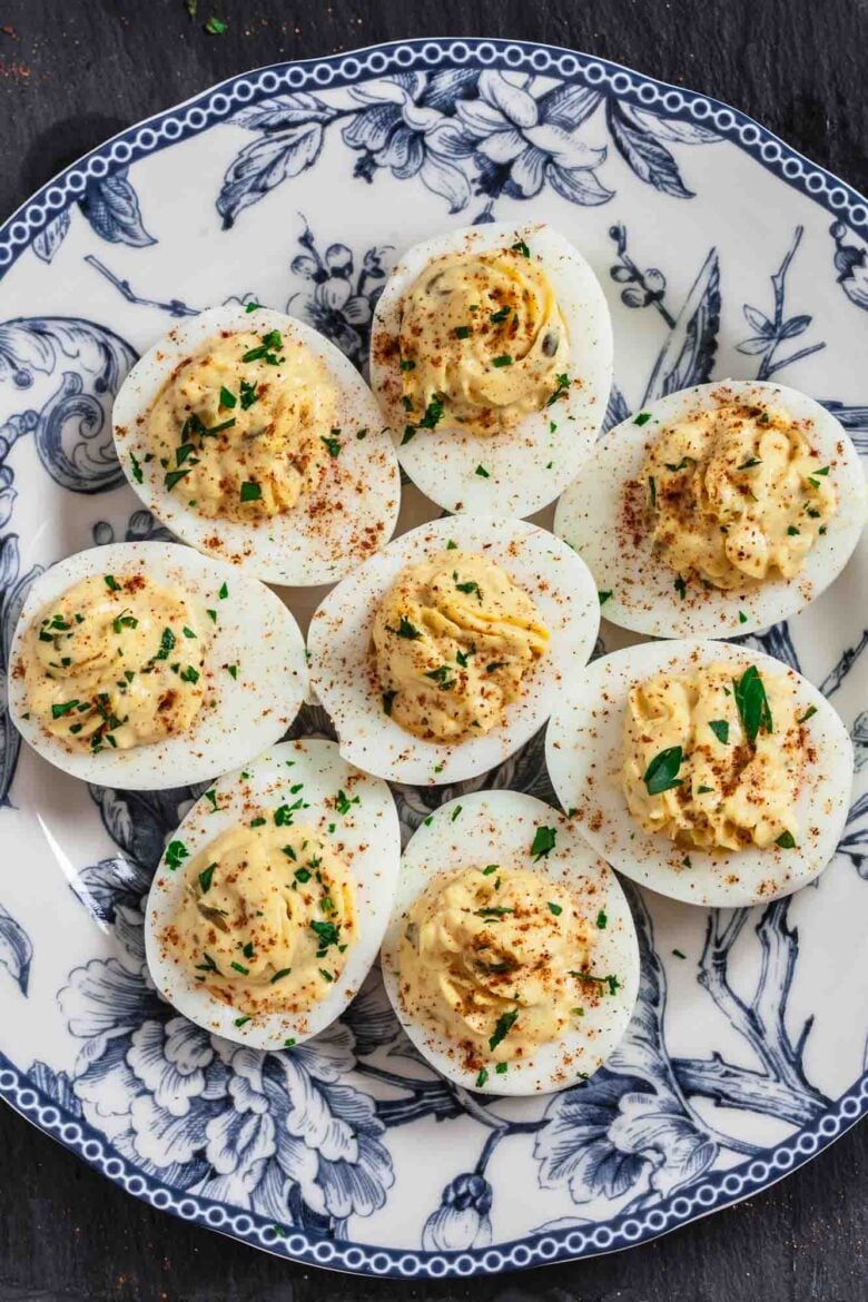 A plate with eight creamy deviled eggs, topped with paprika and chopped herbs, arranged neatly on a decorative blue and white dish—an ideal make-ahead appetizer for any gathering.