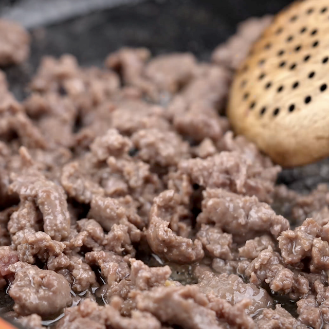 Close-up of sausage being cooked in a pan with a slotted wooden spatula.