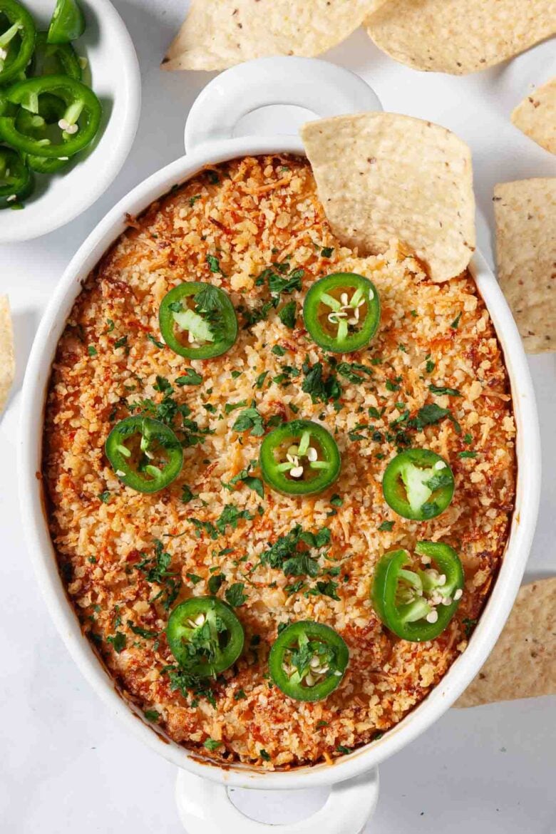 An oval baking dish filled with Jalapeño Popper Party Dip, topped with golden breadcrumbs, jalapeño slices, and chopped herbs, surrounded by tortilla chips.