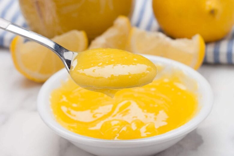 A spoon holds a serving of homemade lemon curd above a small white bowl filled with more curd; sliced lemons and a striped cloth are in the background.