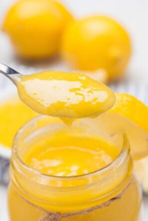 A spoonful of homemade lemon curd held above an open glass jar, with fresh lemons in the background.