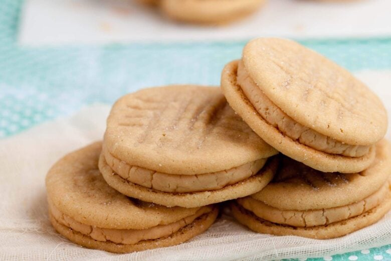 A stack of Peanut Butter Sandwich Cookies with a buttery, creamy peanut butter filling sits on a light cloth, with more cookies blurred in the background.