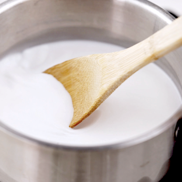 A wooden spoon stirring a white liquid in a stainless steel pot, preparing creamy Coconut Rice.