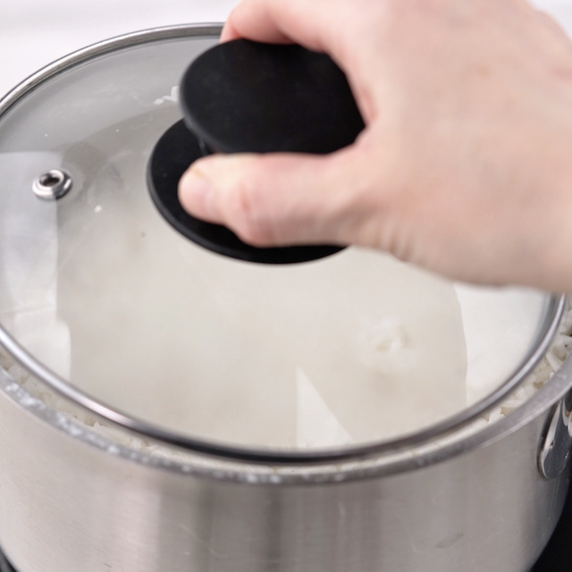 A hand is placing a glass lid onto a stainless steel pot that appears to be cooking Coconut Rice, a classic Asian style dish.