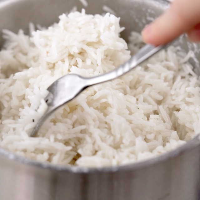 A hand uses a fork to fluff coconut rice in a stainless steel pot, preparing this tropical side dish to perfection.