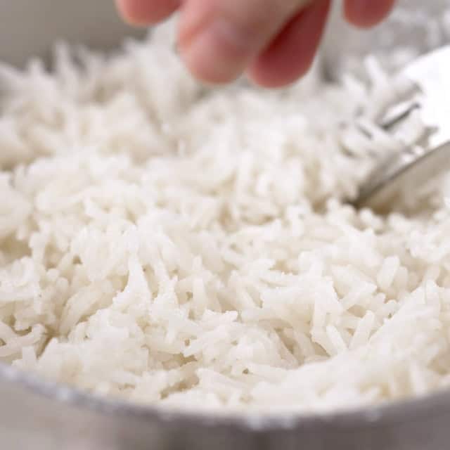 A hand seasons cooked Asian style Coconut Rice in a bowl with a fork, creating the perfect side dish.