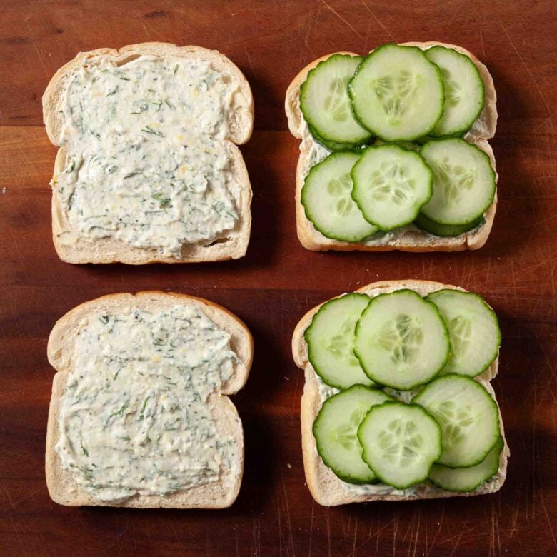 Cucumber sandwich bread slices being assembled with cream cheese spread and sliced cucumbers.