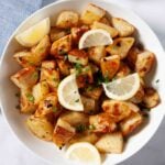 A bowl of Greek lemon potatoes roasted with olive oil and garlic, garnished with chopped chives and lemon wedges, with a small dish of lemon slices in the background.