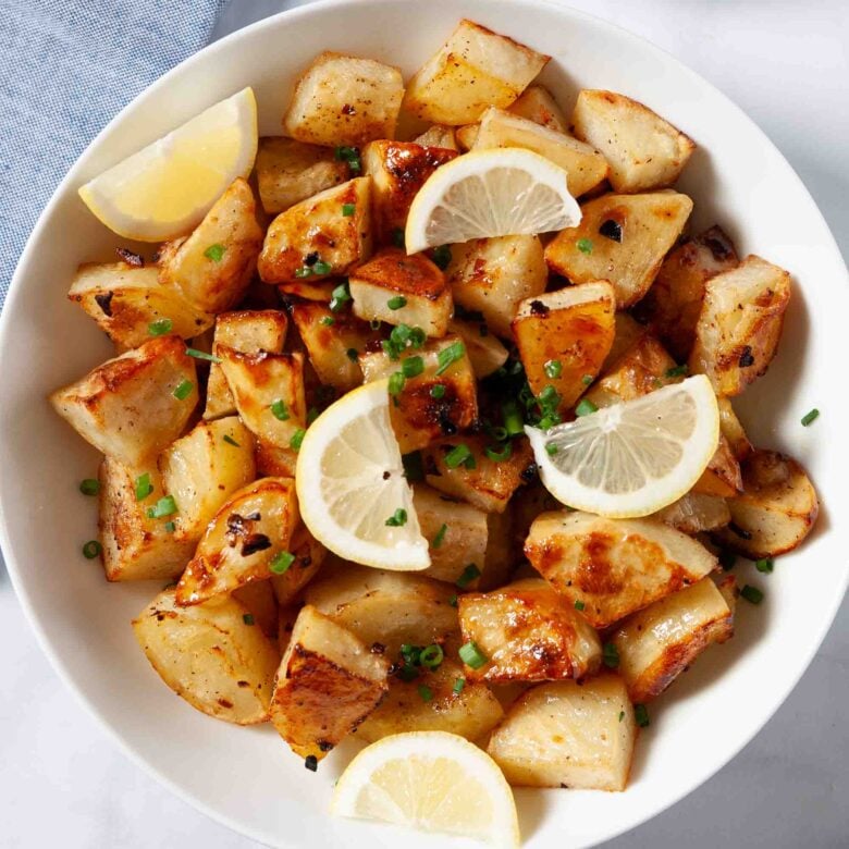 A bowl of Greek lemon potatoes roasted with olive oil and garlic, garnished with chopped chives and lemon wedges, with a small dish of lemon slices in the background.