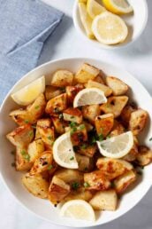 A bowl of Greek lemon potatoes roasted with olive oil and garlic, garnished with chopped chives and lemon wedges, with a small dish of lemon slices in the background.