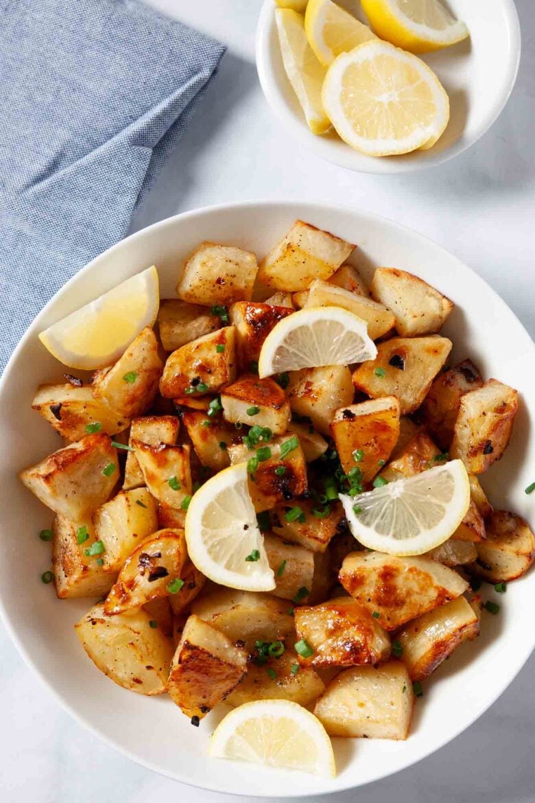 A bowl of Greek lemon potatoes roasted with olive oil and garlic, garnished with chopped chives and lemon wedges, with a small dish of lemon slices in the background.