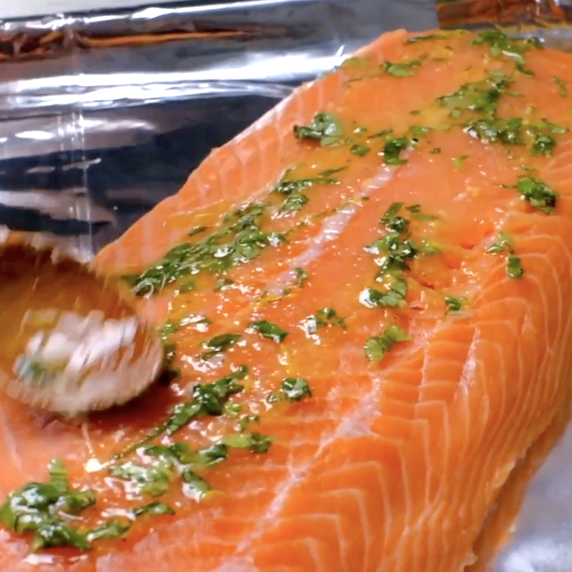 A wooden spoon spreads a herb and oil marinade over a raw salmon fillet on a sheet of aluminum foil.
