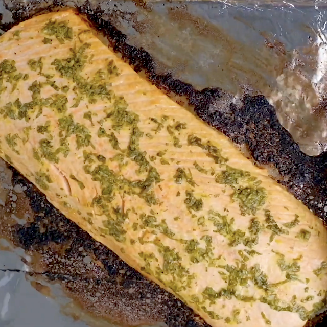 A wooden spoon spreads a herb and oil marinade over a raw salmon fillet on a sheet of aluminum foil.
