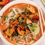 A bowl of Thai curry noodles with chicken, vegetables, fresh herbs, and lime slices in a reddish broth, served with chopsticks.