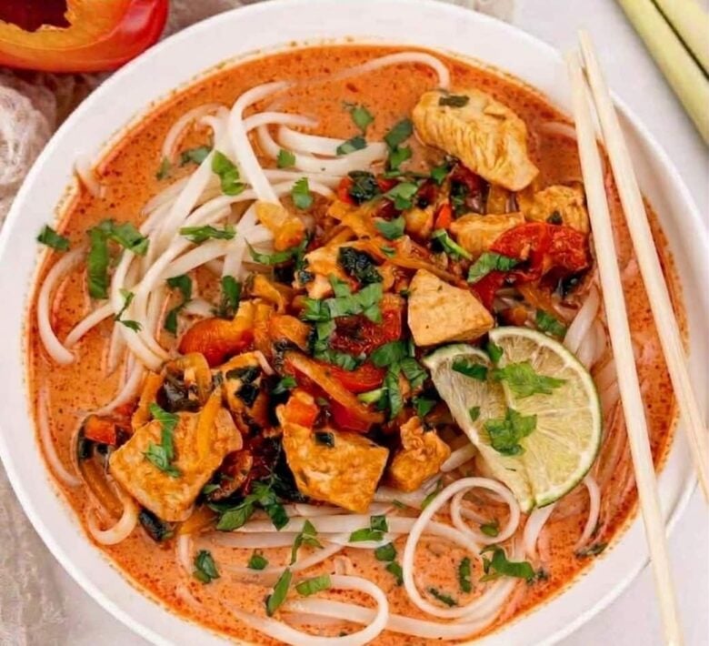 A bowl of Thai curry noodles with chicken, vegetables, fresh herbs, and lime slices in a reddish broth, served with chopsticks.