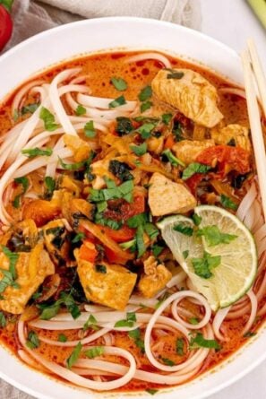 A bowl of Thai curry noodles with chicken, vegetables, fresh herbs, and lime slices in a reddish broth, served with chopsticks.