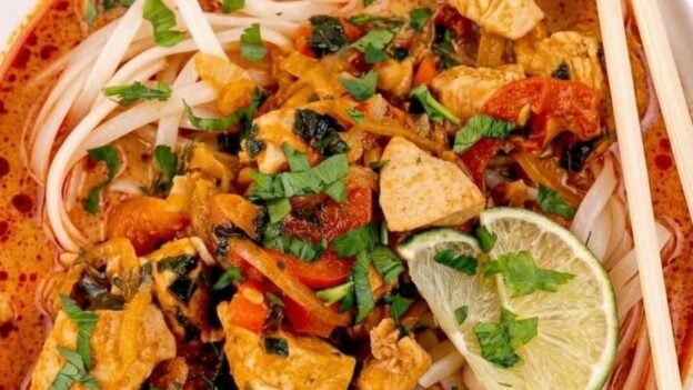 A bowl of Thai curry noodles with chicken, vegetables, fresh herbs, and lime slices in a reddish broth, served with chopsticks.
