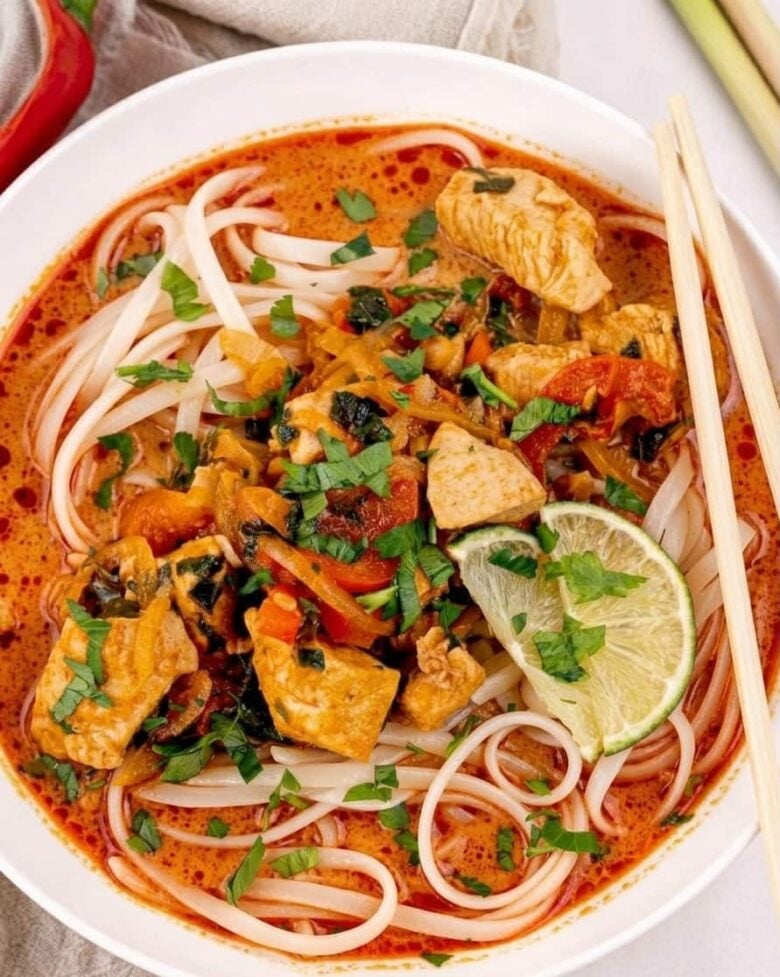 A bowl of Thai curry noodles with chicken, vegetables, fresh herbs, and lime slices in a reddish broth, served with chopsticks.