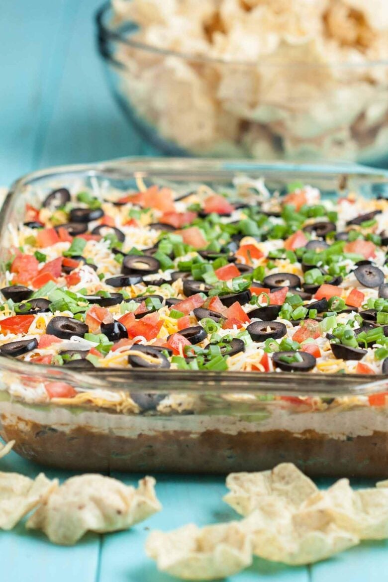 Vertical side shot of 7 layer Mexican dip in a glass serving dish, along with scoop tortilla chips. 