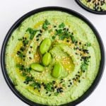 Edamame hummus garnished with edamame beans and herbs and sesame seeds in a black bowl.