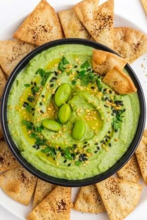 Edamame Hummus in a black bowl with various chips surrounding it.