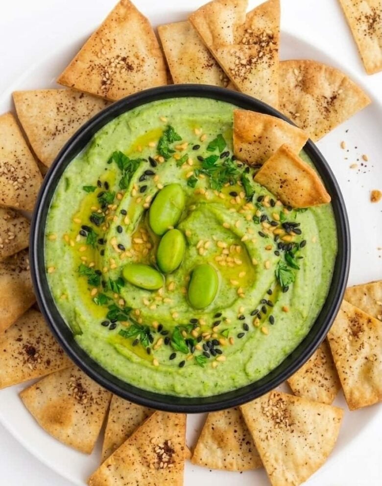 Edamame Hummus in a black bowl with various chips surrounding it.