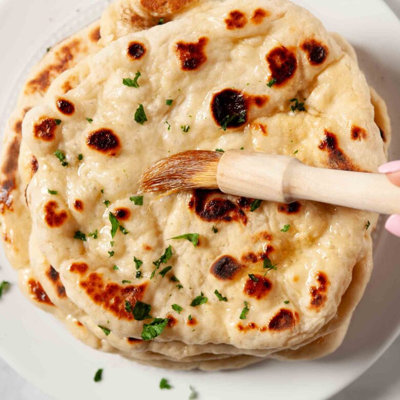Hand brushing melted garlic butter and fresh parsley onto a golden, bubbly naan bread in a cast iron skillet.