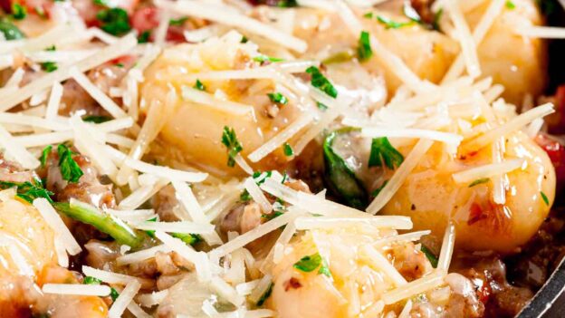 Closeup shot of a bowl of gnocchi with sausage topped with cheese and fresh herbs.