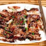 Grilled Teriyaki Chicken sliced on a bed of rice on white plate, garnished with green onions and sesame seeds.