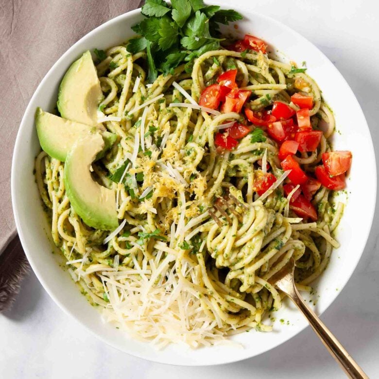 Bowl of creamy avocado pasta with green pesto sauce, topped with shredded cheese, chopped tomatoes, avocado slices, and parsley.
