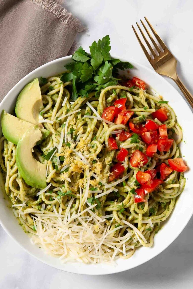 Bowl of creamy avocado pasta with green pesto sauce, topped with shredded cheese, chopped tomatoes, avocado slices, and parsley.