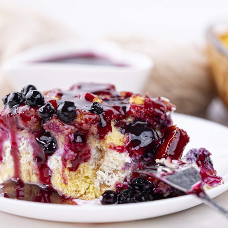 A close-up of a square slice of Blueberry French Toast Casserole topped with blueberries and purple syrup, served on a white plate.