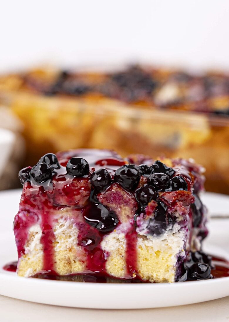 A close-up of a square slice of Blueberry French Toast Casserole topped with blueberries and purple syrup, served on a white plate.