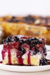 A close-up of a square slice of Blueberry French Toast Casserole topped with blueberries and purple syrup, served on a white plate.
