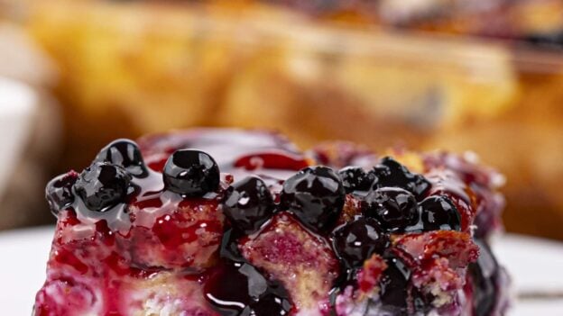 A close-up of a square slice of Blueberry French Toast Casserole topped with blueberries and purple syrup, served on a white plate.