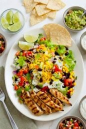 A homemade Chipotle Chicken Burrito Bowl featuring grilled chicken, lettuce, black beans, corn, tomatoes, cheese, rice, and tortilla chips, surrounded by lime wedges, guacamole, salsa, and sour cream in small bowls.