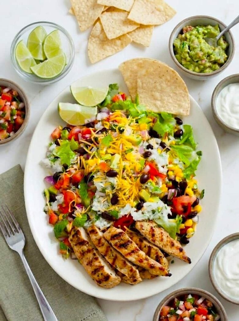 A homemade Chipotle Chicken Burrito Bowl featuring grilled chicken, lettuce, black beans, corn, tomatoes, cheese, rice, and tortilla chips, surrounded by lime wedges, guacamole, salsa, and sour cream in small bowls.