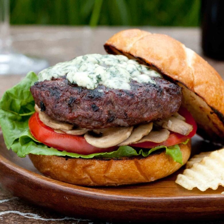 A gourmet burger with lettuce, tomato, mushrooms, and a grilled beef patty topped with melted blue cheese on a toasted bun, set on a wooden plate.