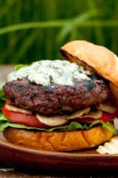 A gourmet burger with lettuce, tomato, mushrooms, and a grilled beef patty topped with melted blue cheese on a toasted bun, set on a wooden plate.