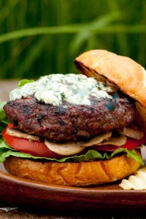 A gourmet burger with lettuce, tomato, mushrooms, and a grilled beef patty topped with melted blue cheese on a toasted bun, set on a wooden plate.