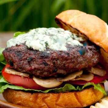 A gourmet burger with lettuce, tomato, mushrooms, and a grilled beef patty topped with melted blue cheese on a toasted bun, set on a wooden plate.