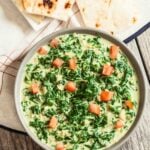 A bowl of Indian Creamed Spinach garnished with diced tomatoes, served with pieces of naan on a cloth.