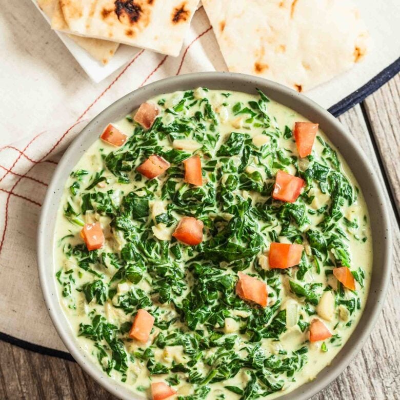 A bowl of Indian Creamed Spinach garnished with diced tomatoes, served with pieces of naan on a cloth.